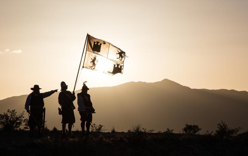 El Camino Real de Tierra Adentro National Historic Trail in New Mexico. Three men dressed as Spanish explorers wave a flag as they stand on a hillside in bright sunlight.