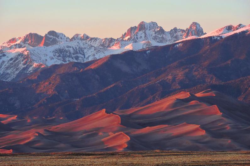 Tall sand dunes and mountains glow in the red light of sunset.