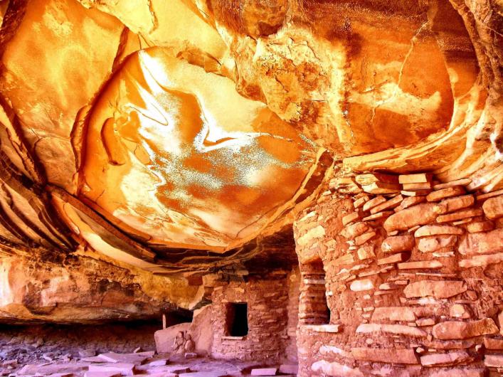 The ruins of old stone buildings are tucked under a sandstone cliff.