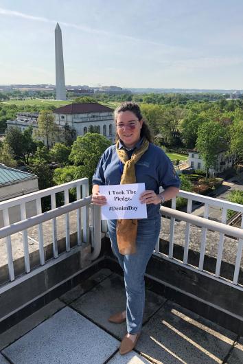 Jennifer Flynn from NPS, outside, standing on the MIB's roof, Washington Monument on the back, holding the I took the Pledge #DenimDay sign. At his back, wildlife set up exhibit item