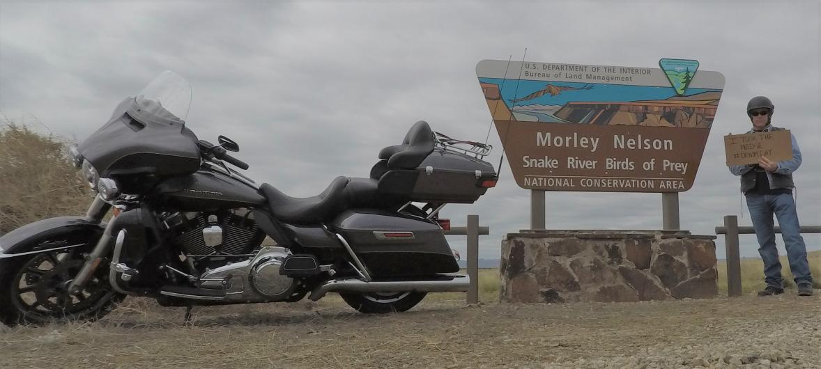Kevin Kirchmeier from Ole holding the I took the Pledge #DenimDay sign, standing by the Morley Nelson Snake River Birds of Prey National Conservation Area. Motorcycle parked by sign. 