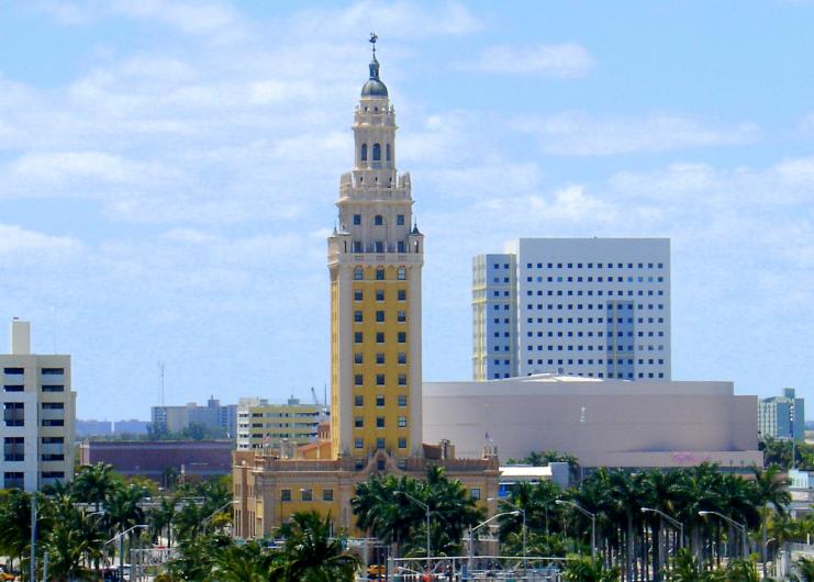 A tall yellow building with an ornate pointed roof stands among the Miami skyline.