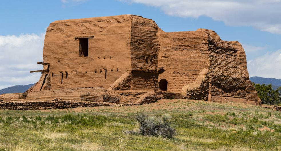 The tall walls of an old brown adobe brick building stand on a grassy hill.