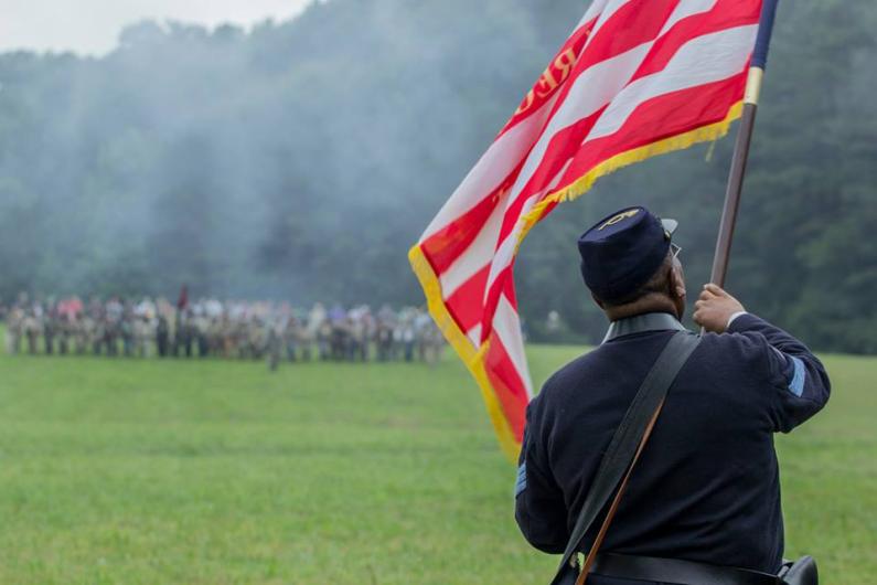 Photo of an African American man wearing a Union Army uniform and waving an American flag in a green field during a battle reenactment.