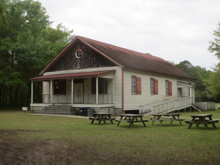 Photo of a long, one story wooden hall with a wide porch and picnic tables on the front lawn.