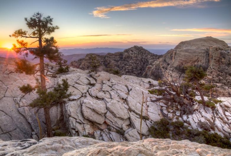 The sun sets over a rocky landscape of mountains and small trees.