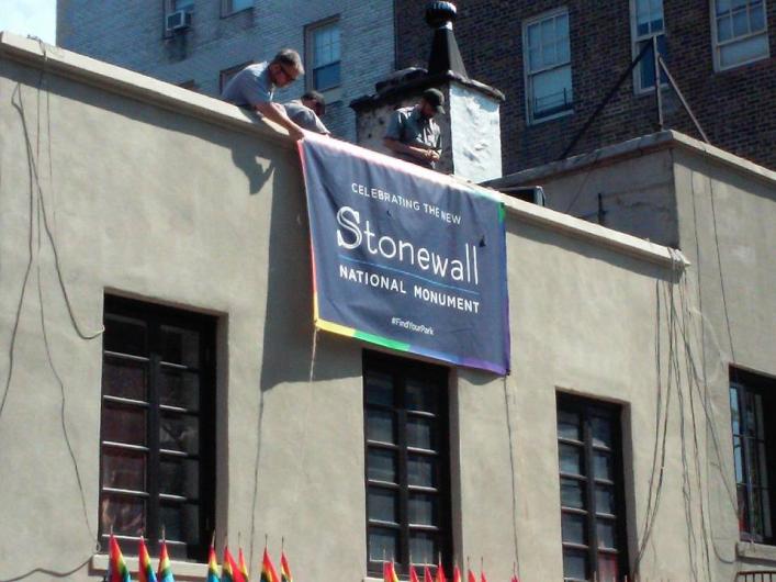 Three National Park Service employees roll down a navy blue banner commemorating the designation of the Stonewall National Monument.