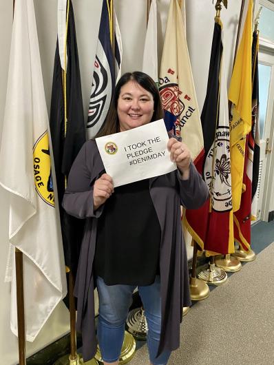 Tara Sweeney from BIA, standing in a hallway,flags at her back, holding the I took the Pledge #DenimDay sign.