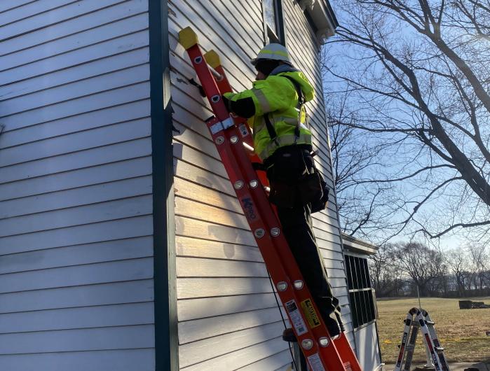 Man wearing a yellow vest and hard hat stands on a ladder propped against white house.​