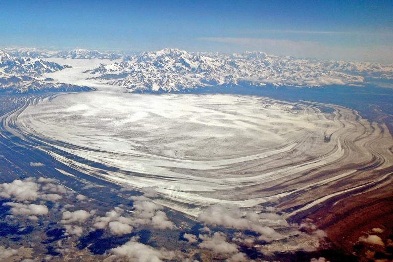 An aerial photo taken from high in the sky shows a huge, flat glacier of ice spreading out from a mountain range.