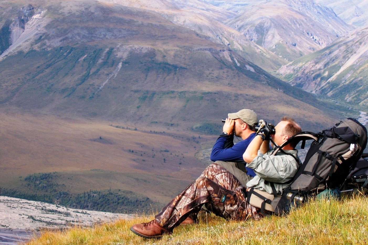 Two people with binoculars sit on a hillside with large backpack.