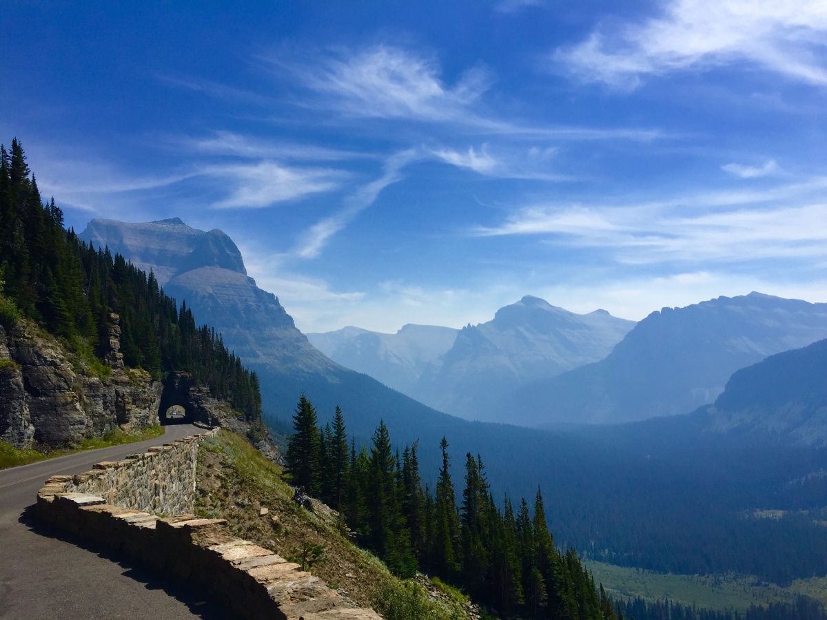 A small road runs along the edge of a tree-filled mountain and leads toward tall mountains far away in the blue sky.