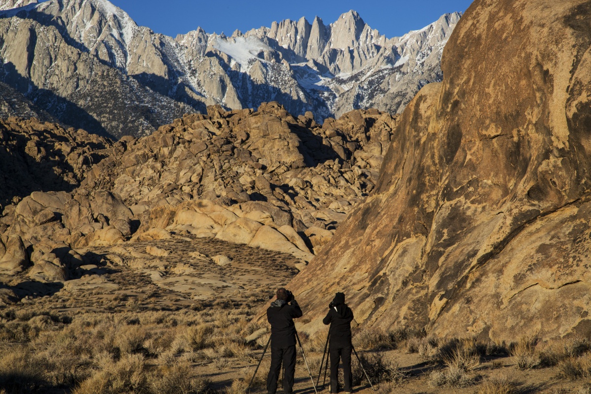 Two photographers in all black point their cameras and tripods toward a tall stretch of rocky brown and grey mountains.