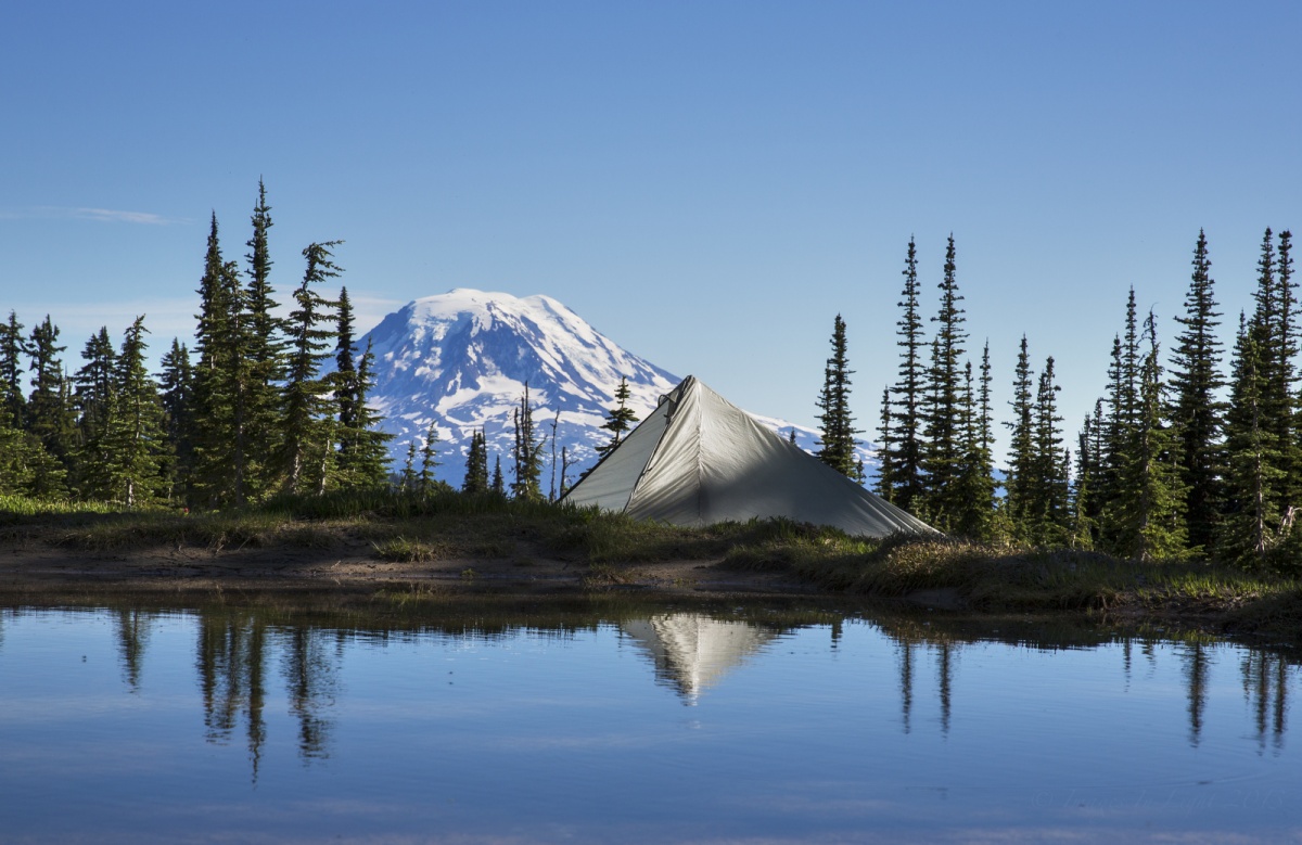 Water reflects a row of tall green trees and a white tent with a snow covered mountain peeking through the back surrounded by bright blue sky.