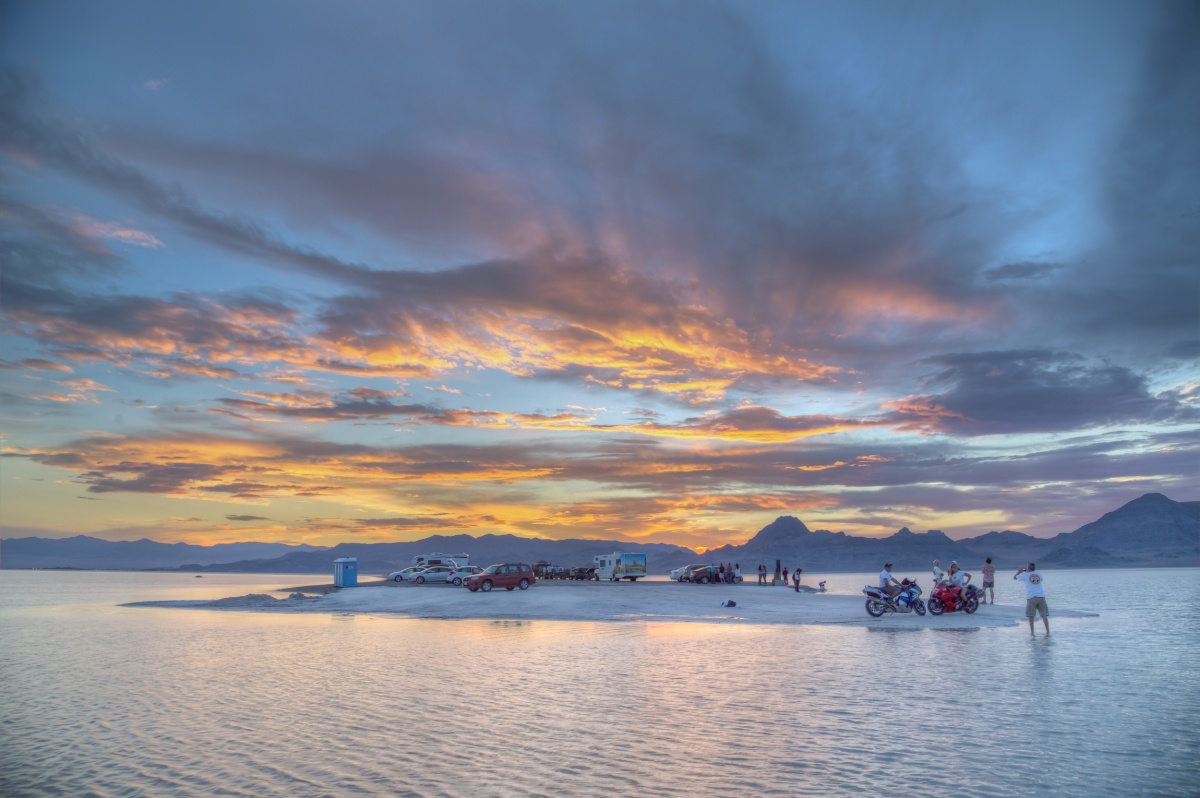 A small island filled with cars and people sits in a light blue water with a gorgeous blue, yellow, and pink sunset filling the sky.