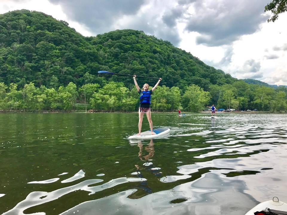 Girl in blue life jacket stands on paddle board in river.