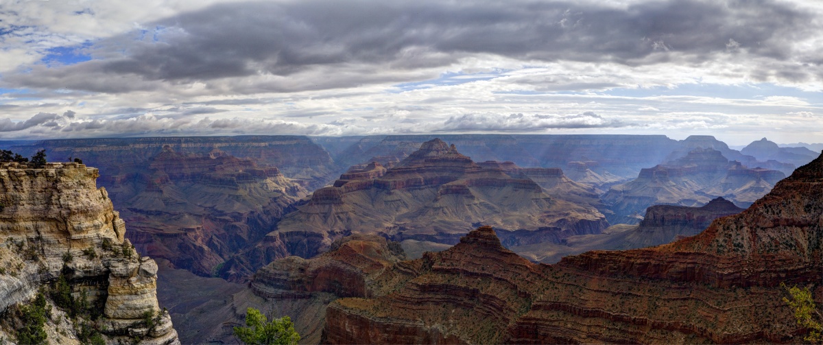 Rock peaks with bands of color sit in the middle of the Grand Canyon. A clouded sky lets in rays of sunshine.