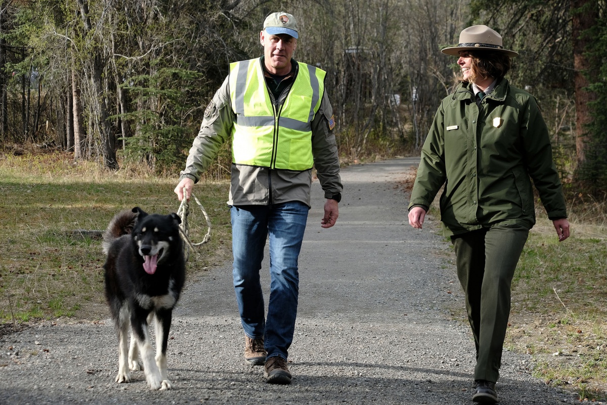 Secretary Zinke and a park ranger smile as they take a sled dog for a walk down a path surrounded by green grass.
