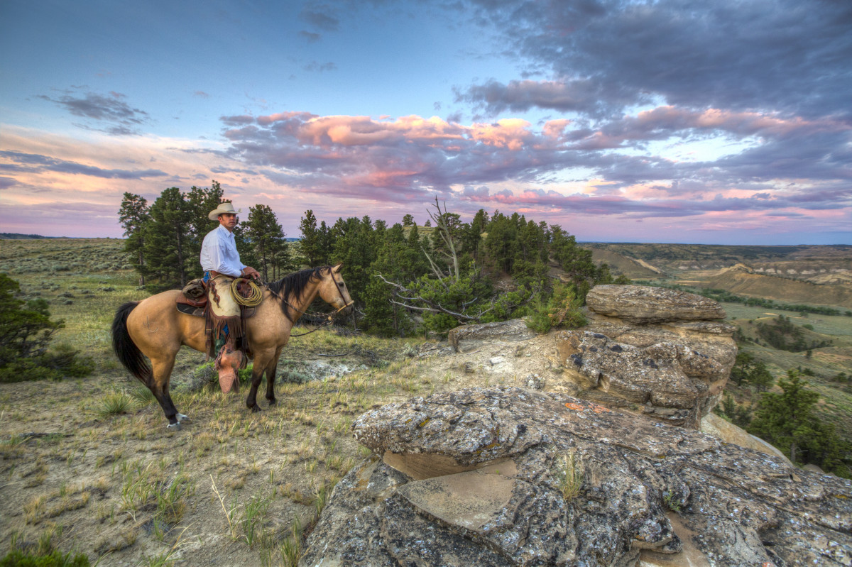 A cowboy on a horse watches from the edge of a cliff overlooking a small valley 
