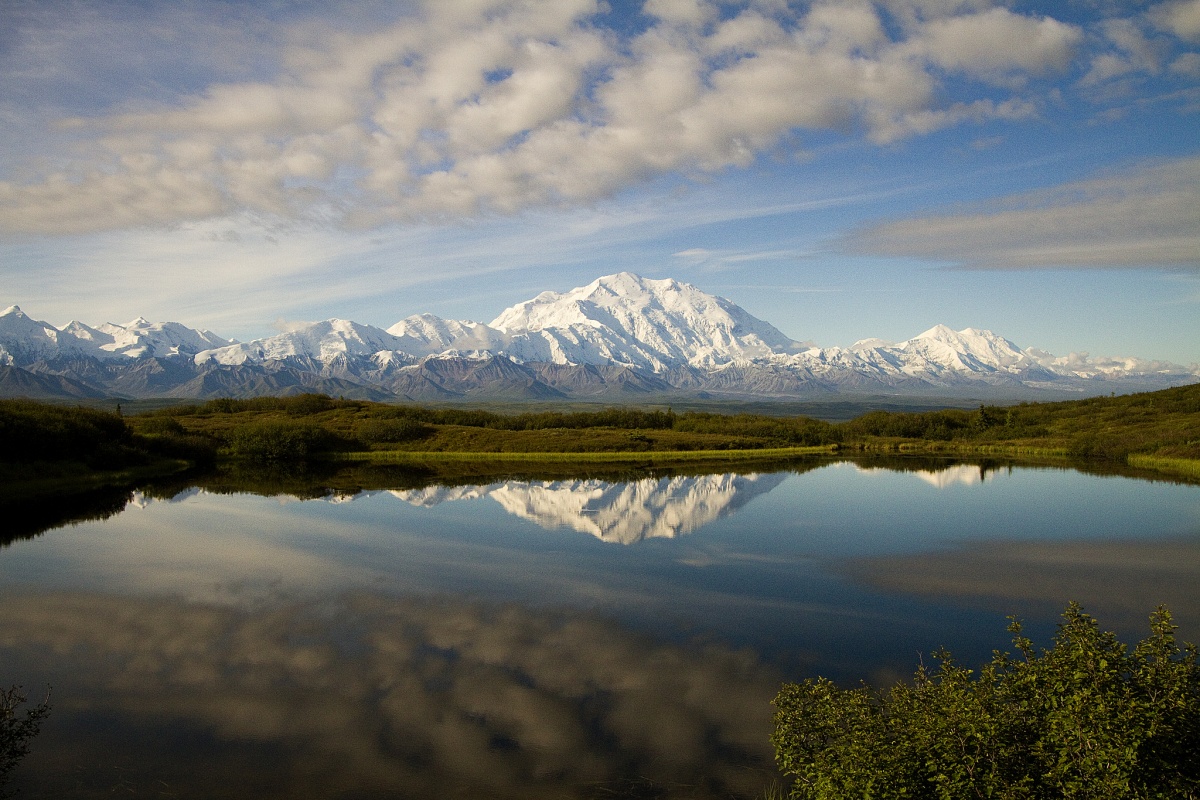 A large clear pond shows the reflection of a white mountain and a bright blue, clouded sky.