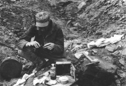 A black and white photo of a man crouching on the ground with tools as he searches for minerals.