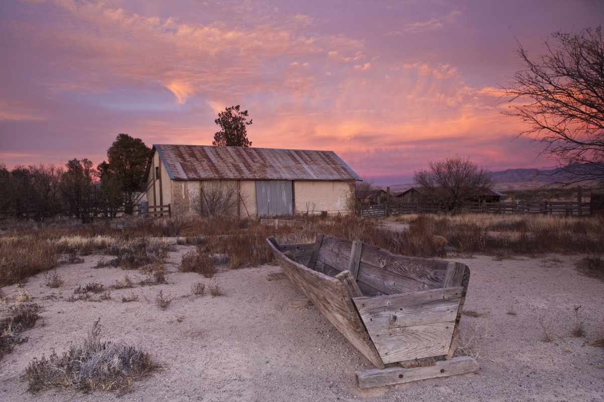 A small ranch house stands among dry, brown and grey field with a warm pink and purple sky behind it.