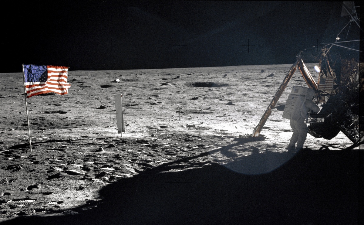 This photo appears black and white, as the lunar surface contrasts against the dark sky. However, on the right side of the photograph, an astronaut climbs into a space ship and on the left side, a bright red, white, and blue American flag flies.