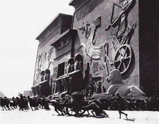 An old black and white photo of the movie set shows people moving a giant monument with lions, four statues of men, and a wall behind them.