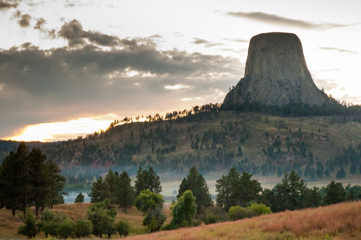 An orange field with green trees is the base for a hill with a tall rock tower stretching into the white clouded sky.