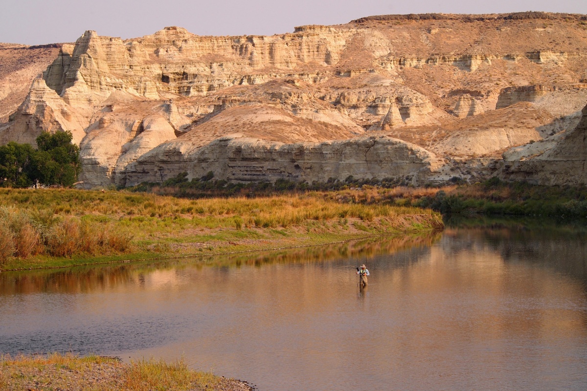A person appears very small as they stand in the middle of a river with surveying equipment, surrounded by green and yellow wildlife and tall, tan rock features in the glow of a sunset.