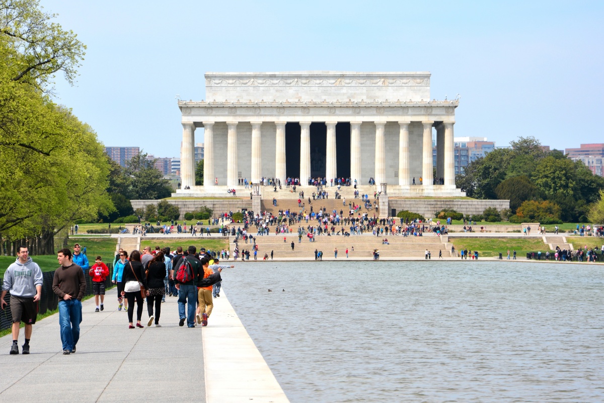 A large white monument stands behind a reflection pool and is surrounded with bright green trees and tons of people walking alongside the pool and monument.