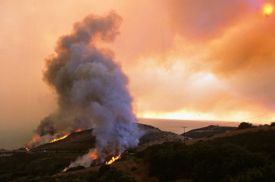 Grey smoke billows into an orange and pink sky from wildfire occurring on brown hilly ground.