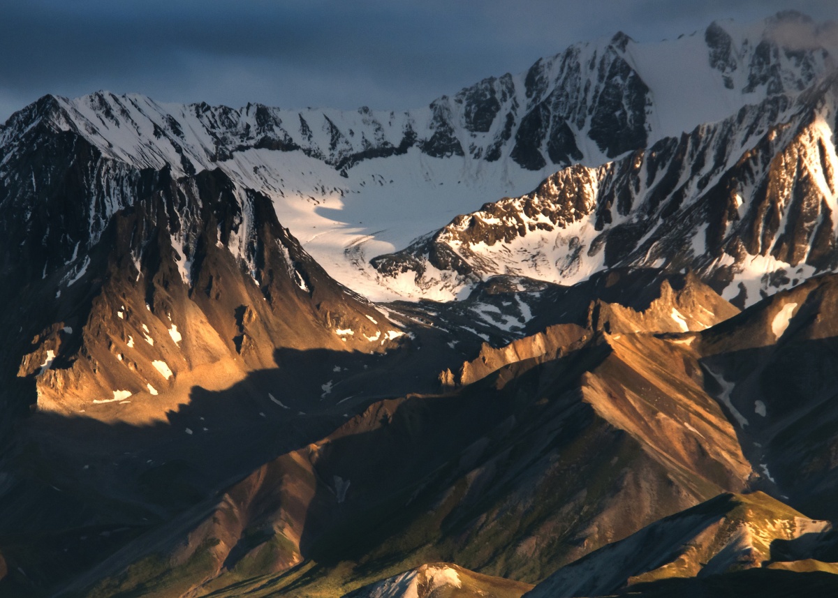 A sunset glows on the side of snow covered brown mountains.