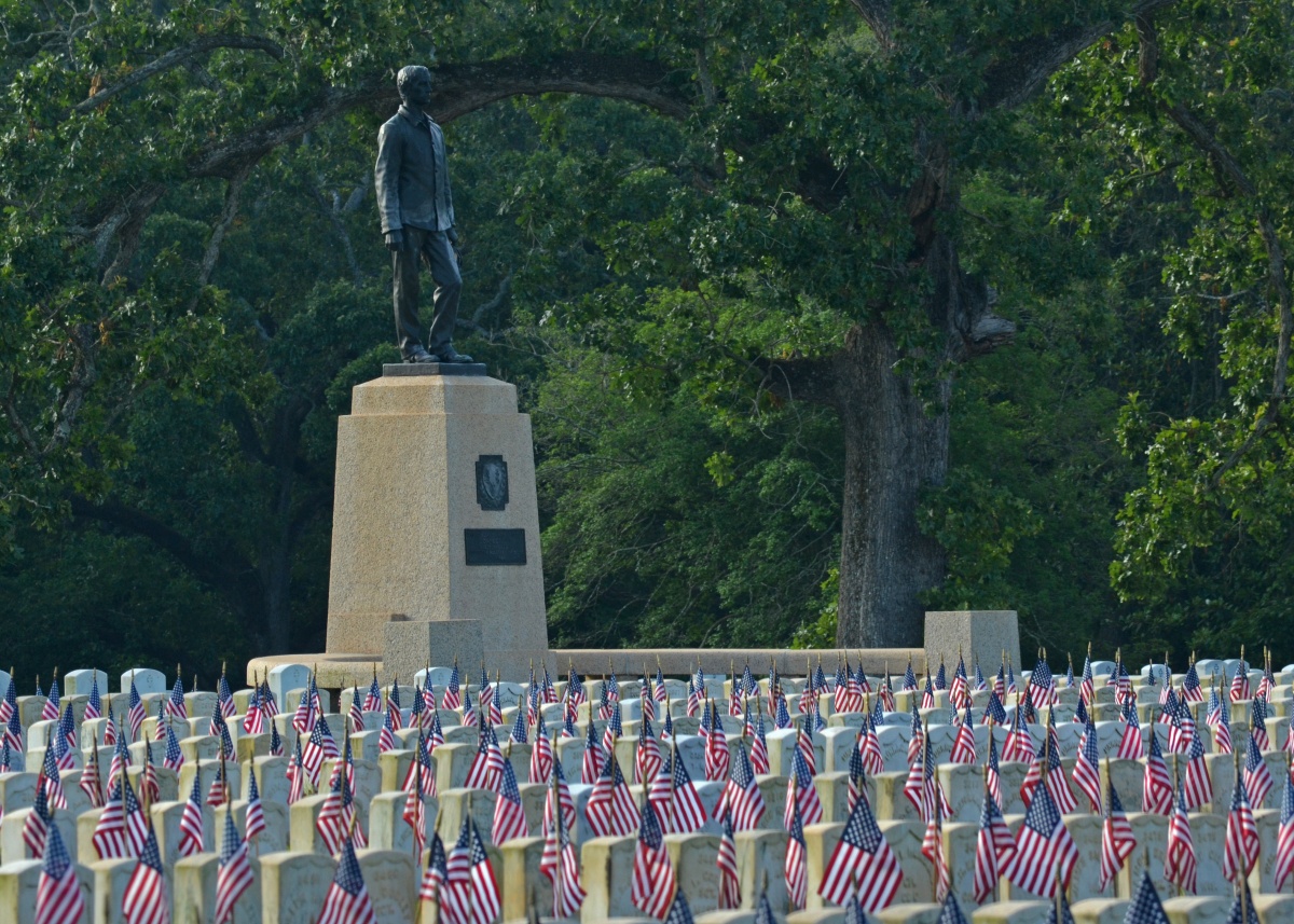 Many small American flags are placed along rows of white gravestones. A tall statue of a man stands against green trees in the background.