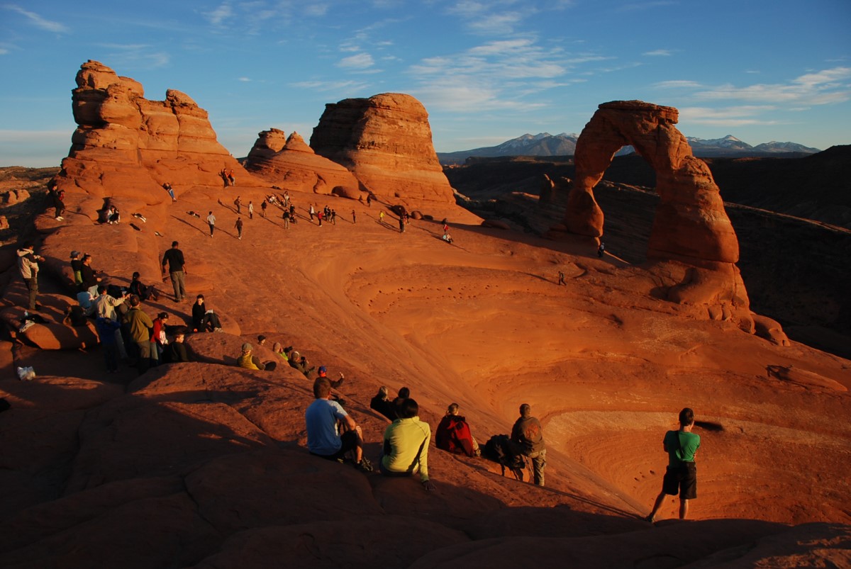 A sparse but wide crowd of visitors circle the orange ridge on the rocky formations of Arches National Park near sunset, embracing the beauty of the endless desert landscape.