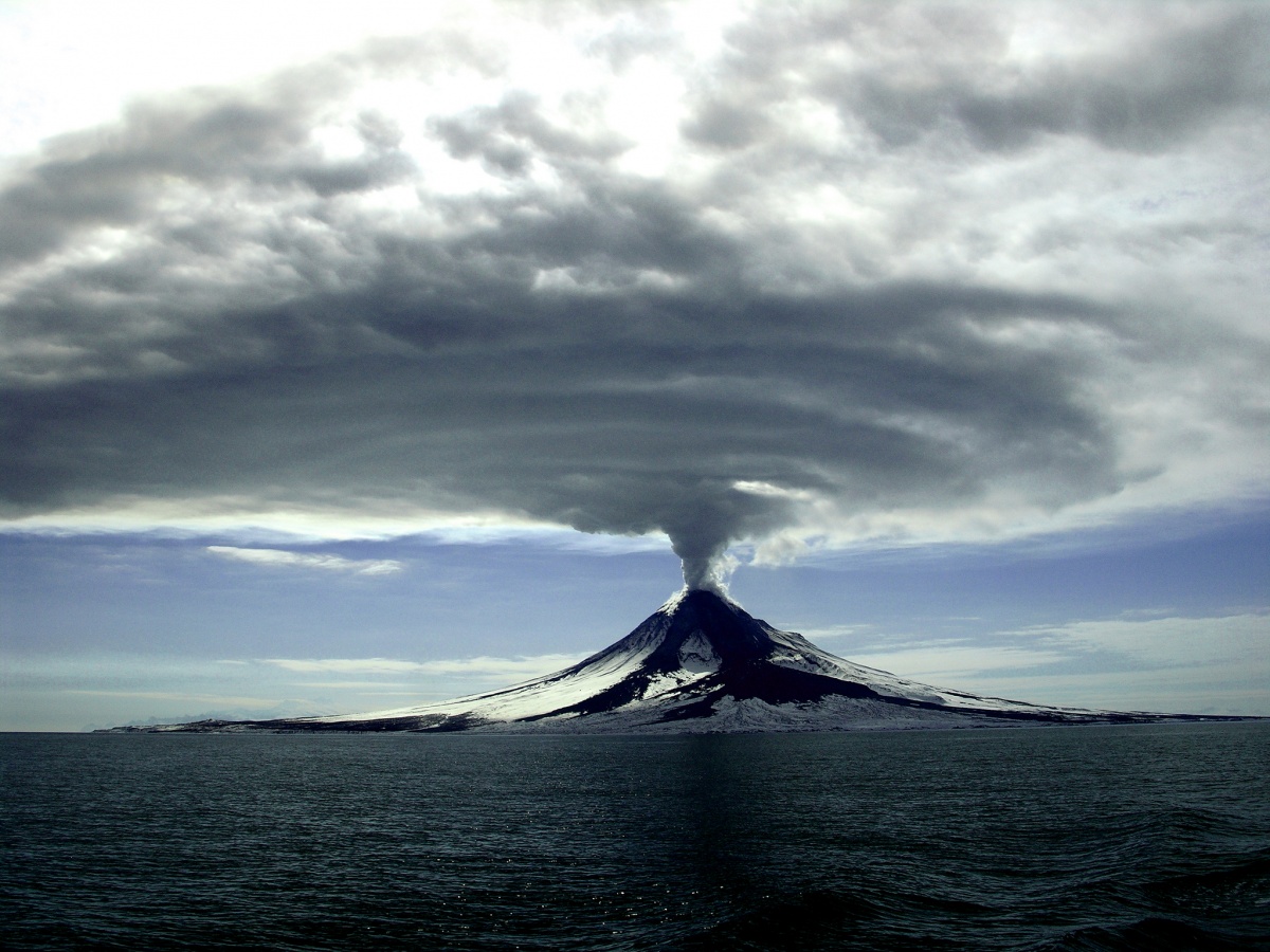 A huge cloud of smoke expands across the sky as it erupts from a cone shaped volcano on the shore of the ocean.