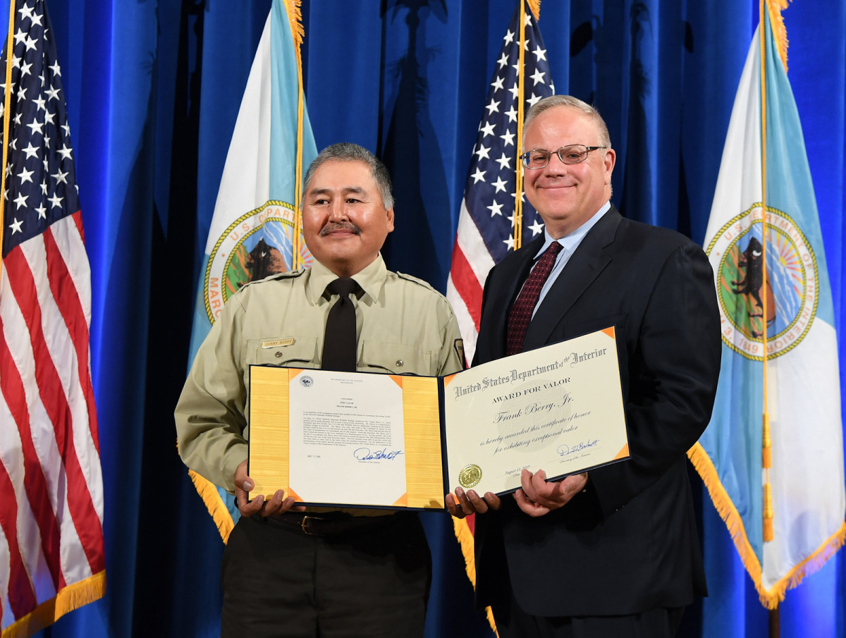 A man in a fish and wildlife service uniform receiving a certificate from Secretary Bernhardt in front of flags and a blue curtain.