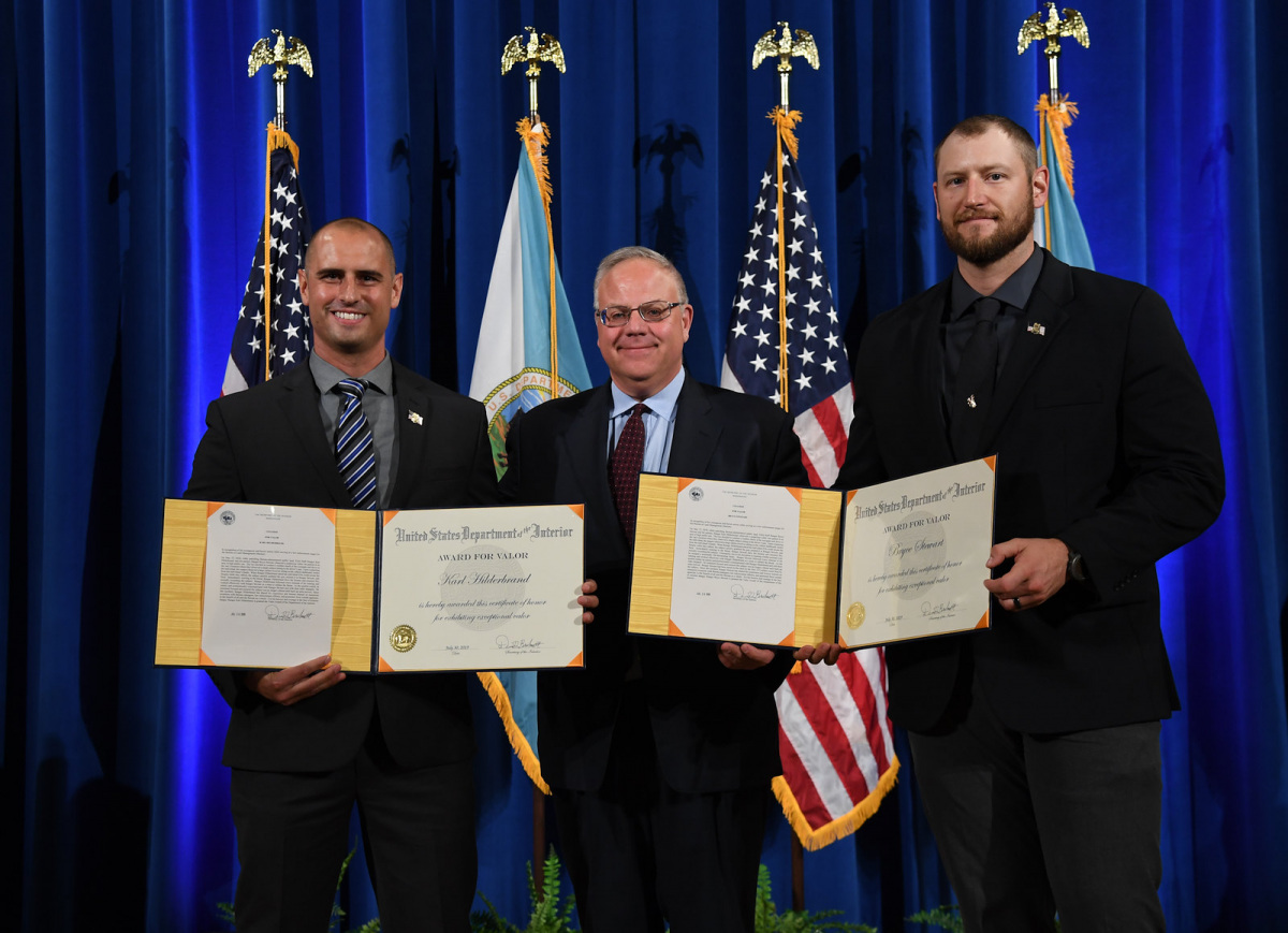 Three white men in suits hold certificates and smile while standing in a line in front of flags and a blue curtain.