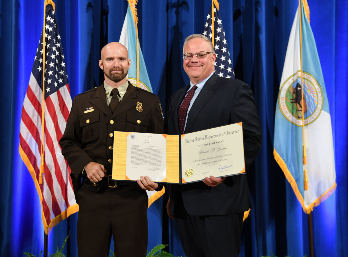 A white man in a brown Fish and Wildlife Service uniform smiles and receives a certificate from a white man in a suit.