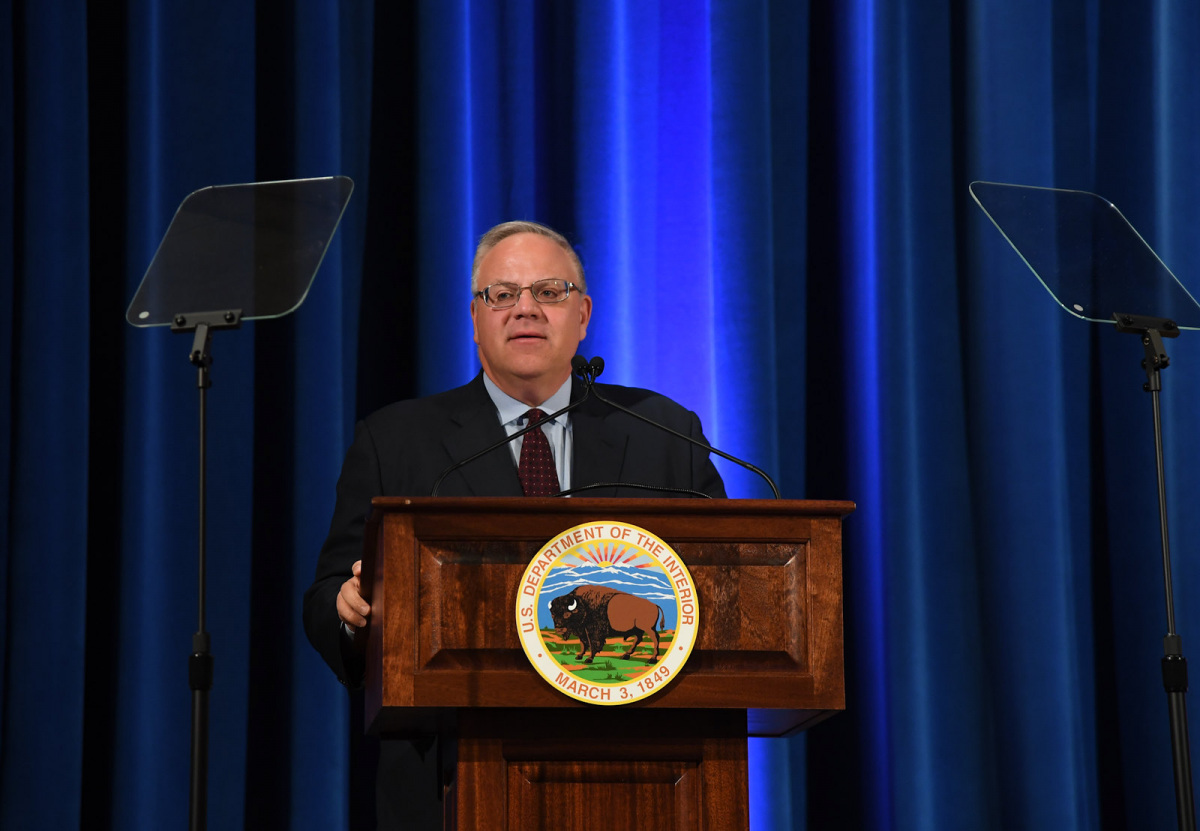 Secretary Bernhardt wears a suit and speaks from a podium with a blue curtain behind him.