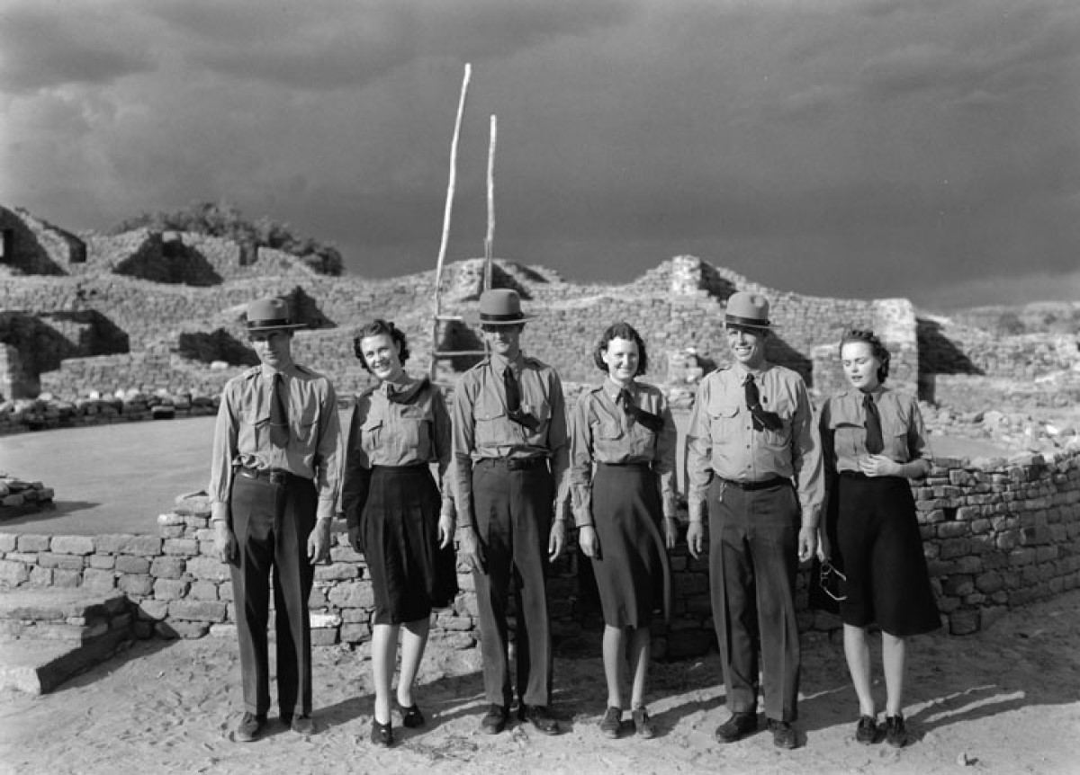 Six youthful rangers in uniform stand on a cloudy, windy day in a lineup in front of sandy brick ruins.