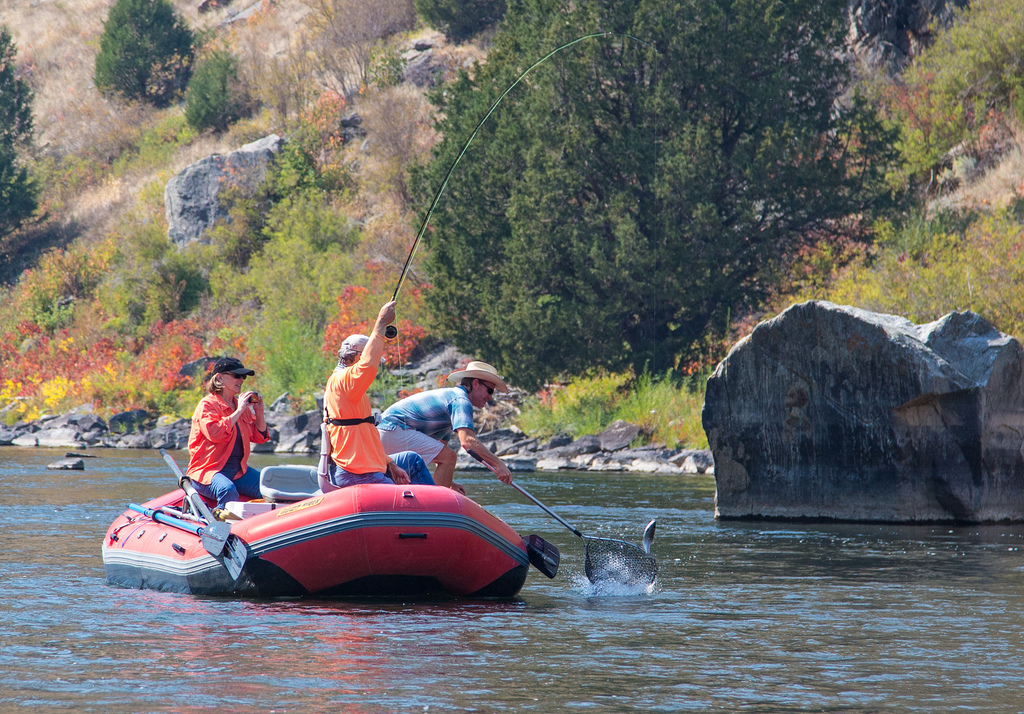 In a red rubber boat floating on a calm river by a grass and tree covered bank, a white man pulls a fish out of the water with his rod and line, while another white man in a hat reaches for the fish with a net as a white woman takes a picture with a camera.