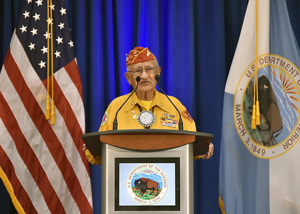 An elderly native american man in a military uniform speaks from a podium with flags and a curtain behind him.