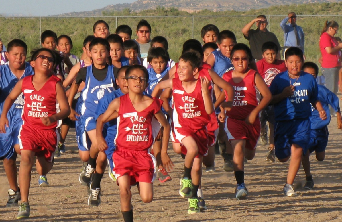 Boys in red and blue athletic clothes run across a dirt field.