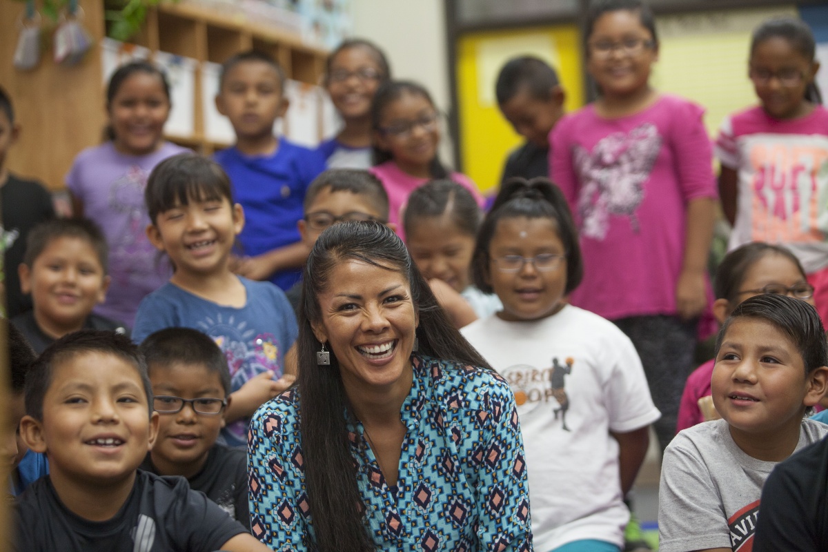 Photo of a classroom with students sitting around their teacher, a native american woman with long brown hair.