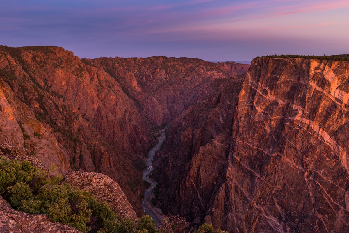 A purple and pink sky glows over tall dark rocks. A river runs through the bottom of the scene.