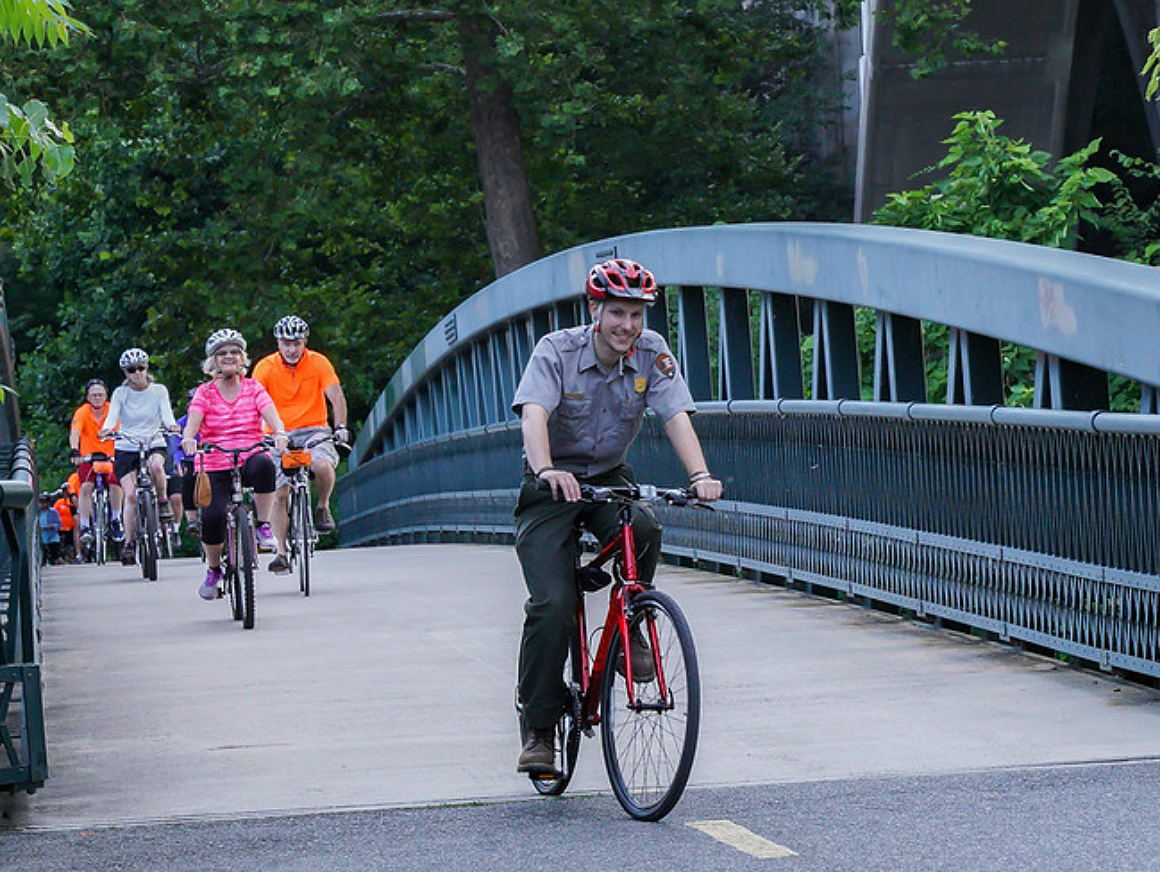 A white male park ranger rides a bike in front of a small group of people also riding bikes across a paved bridge.