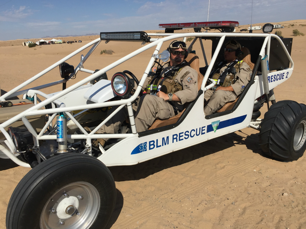 Two men in coveralls and safety gear sit in an open dune buggy parked on a sandy desert plain.