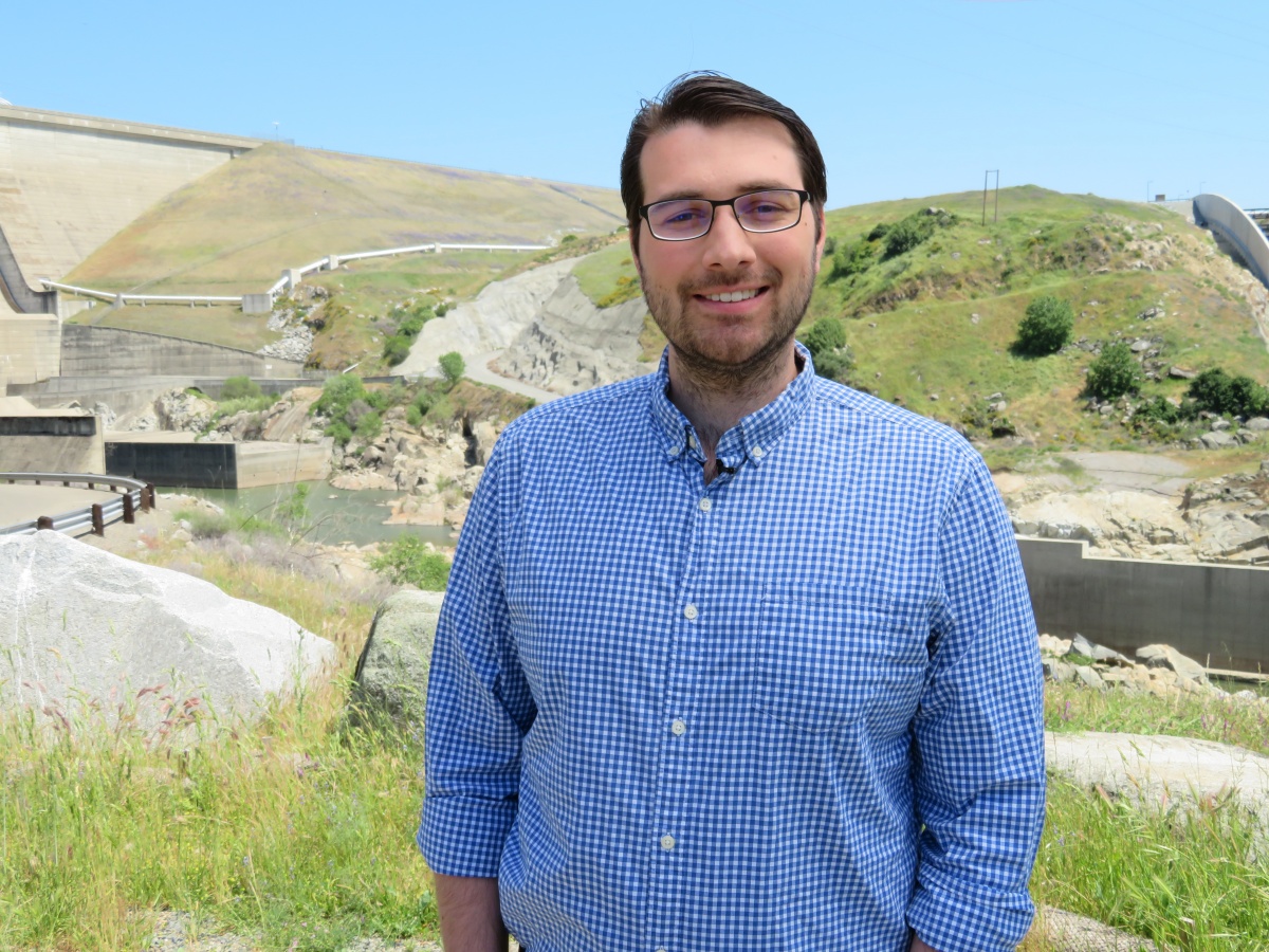 Jim Shannon, a white man with brown hair, a beard and glasses, stands outside in front of a large concrete dam.
