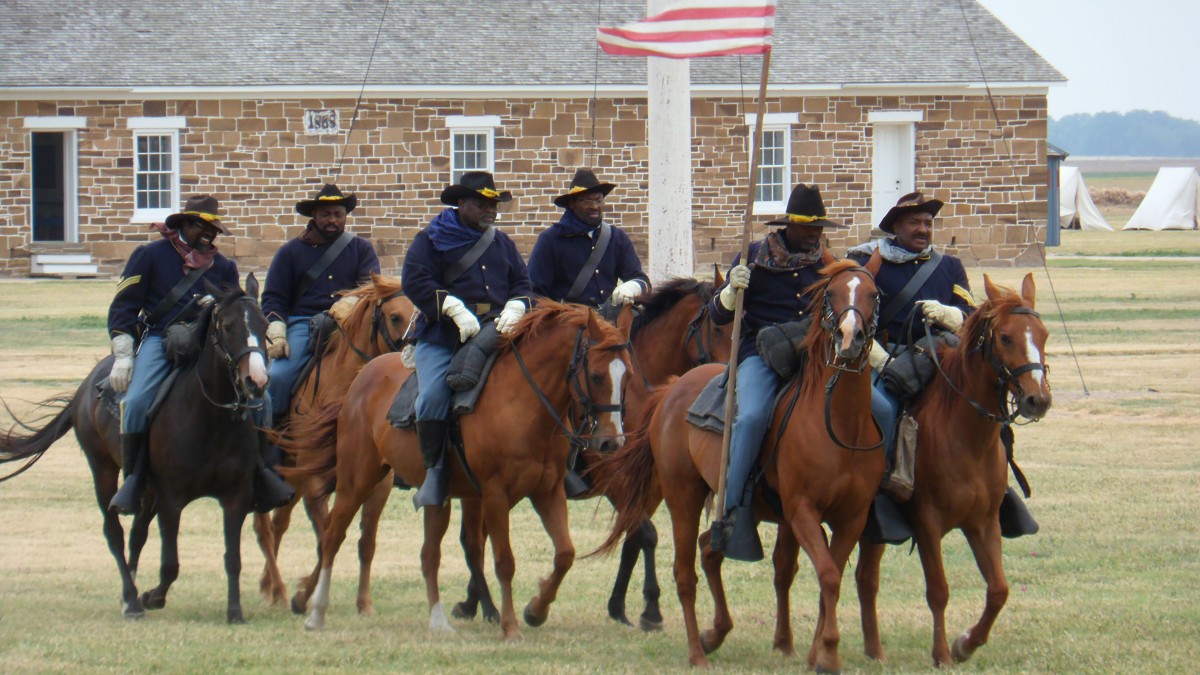 Buffalo Soldier re-enactment group rides horses across green grass.